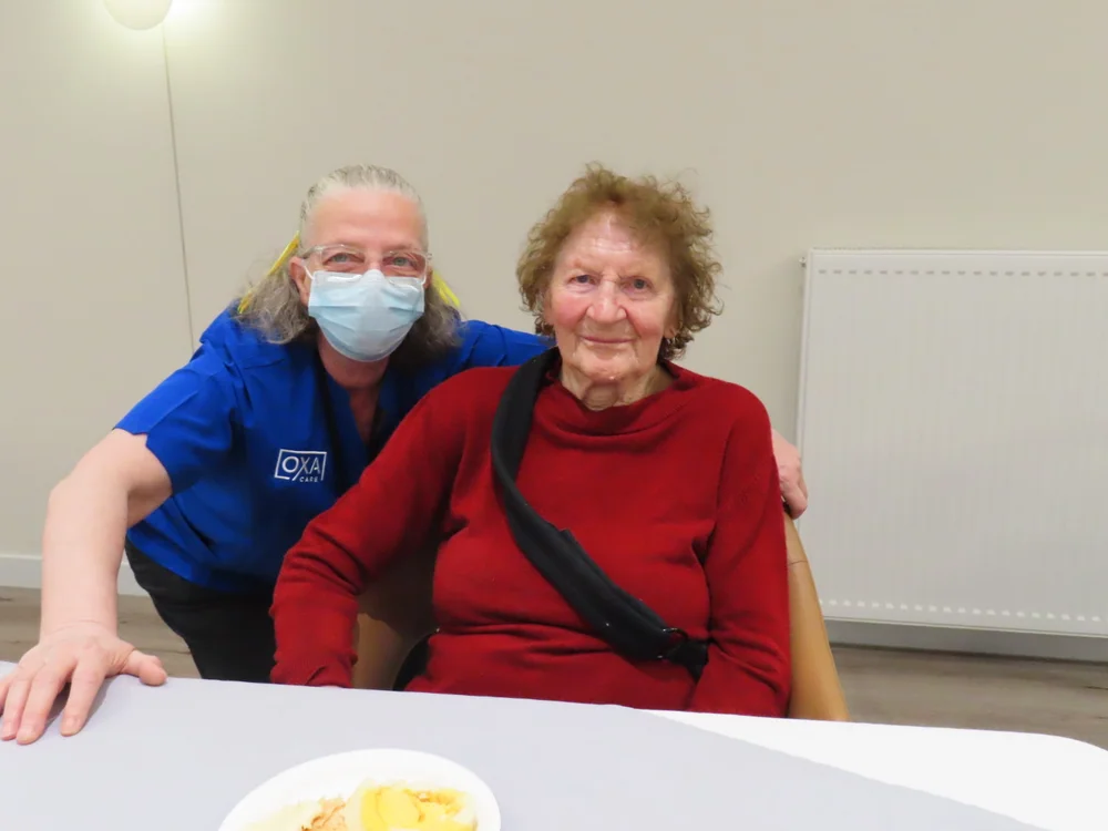 Elderly Russian woman in a red sweater sitting at a table with dessert, smiling beside a caregiver in a blue uniform and mask.