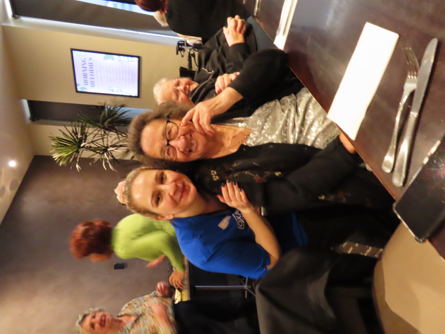 Elderly Russian woman smiling and sitting at a dining table, hugged by a young caregiver in a blue uniform.