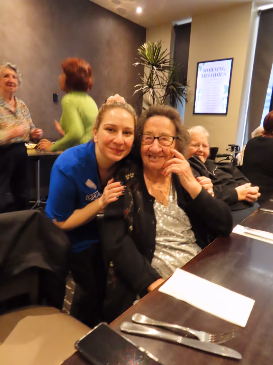 Elderly Russian woman smiling and sitting at a dining table, hugged by a young caregiver in a blue uniform.