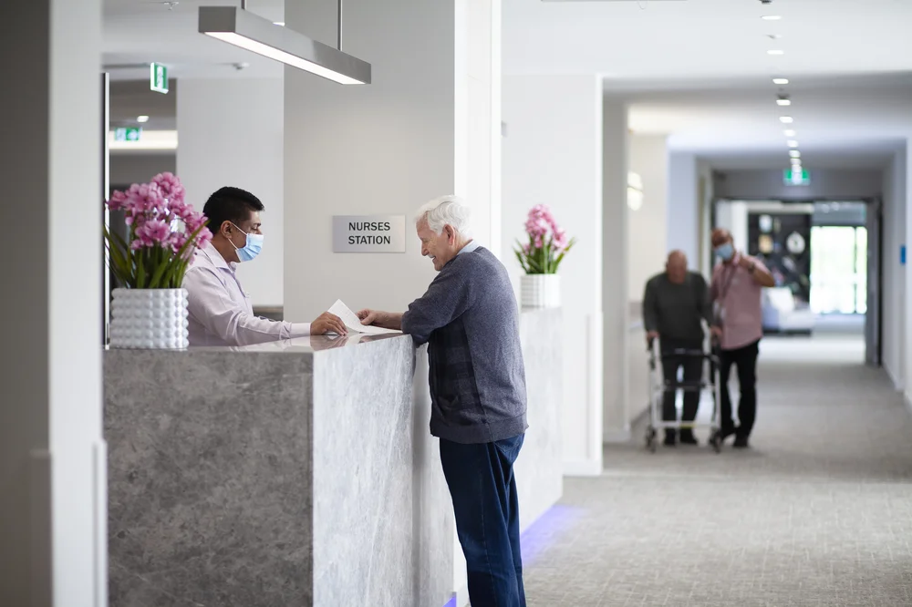 Elderly Serbian man smiling as he speaks with a staff member at the nurses' station.