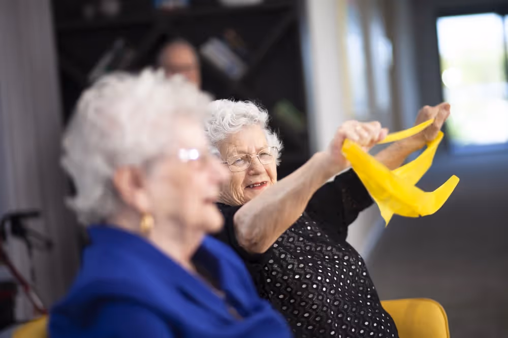 Elderly Serbian woman smiling while exercising with a yellow resistance band during a group activity.