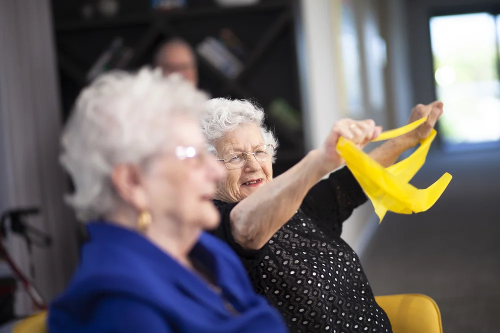 Elderly Serbian woman smiling while exercising with a yellow resistance band during a group activity.