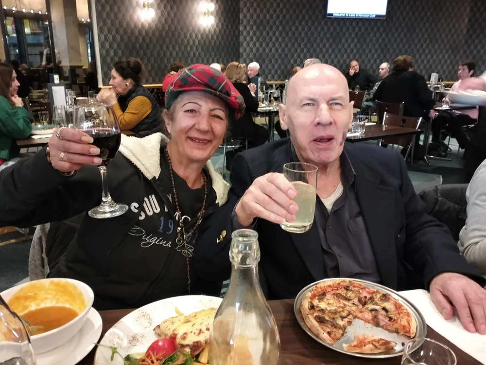 Smiling Serbian couple enjoying a meal at a restaurant, raising glasses of wine and lemonade in a toast.