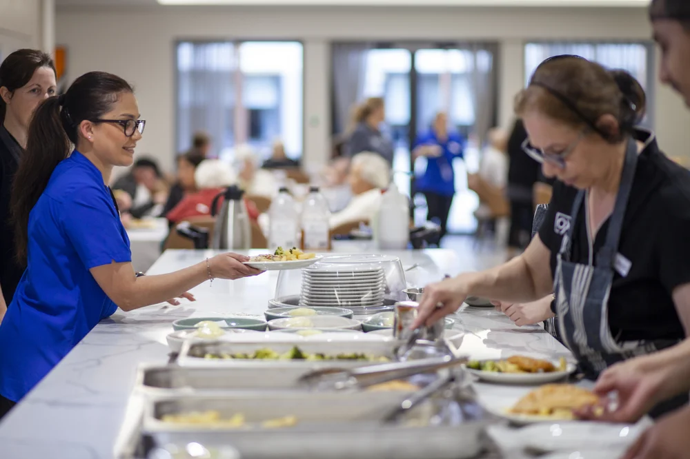 Staff serving meals in a dining area while residents sit and enjoy their food in the background.