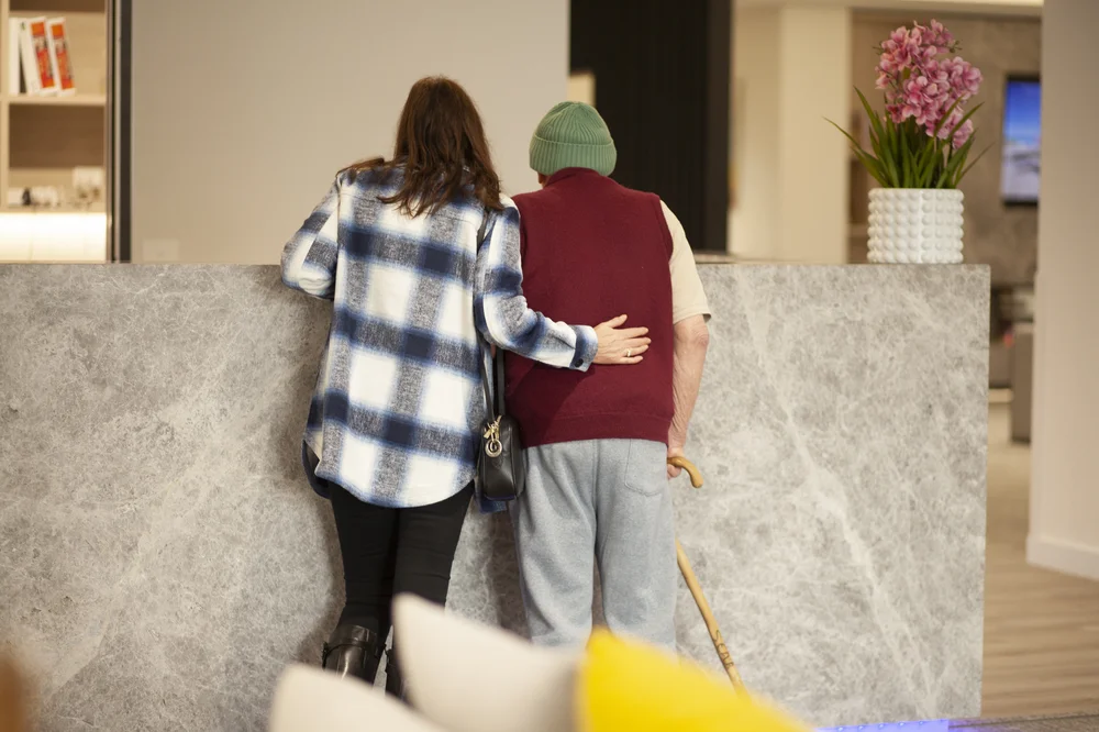 Woman supporting an elderly man with a walking stick as they lean on a reception counter together.