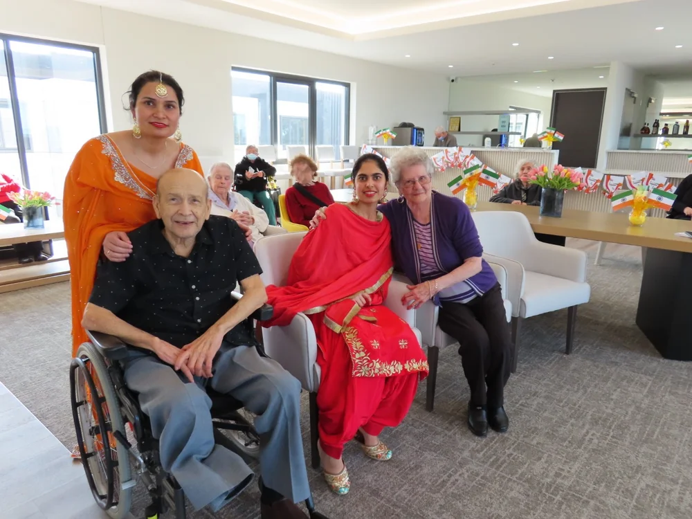 Women in vibrant traditional Indian attire sitting and smiling alongside elderly residents in a decorated common area.