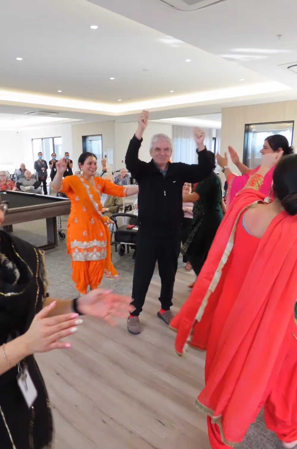 Elderly man joyfully dancing with staff and performers in colourful traditional Indian attire during a lively cultural celebration at an aged care facility.
