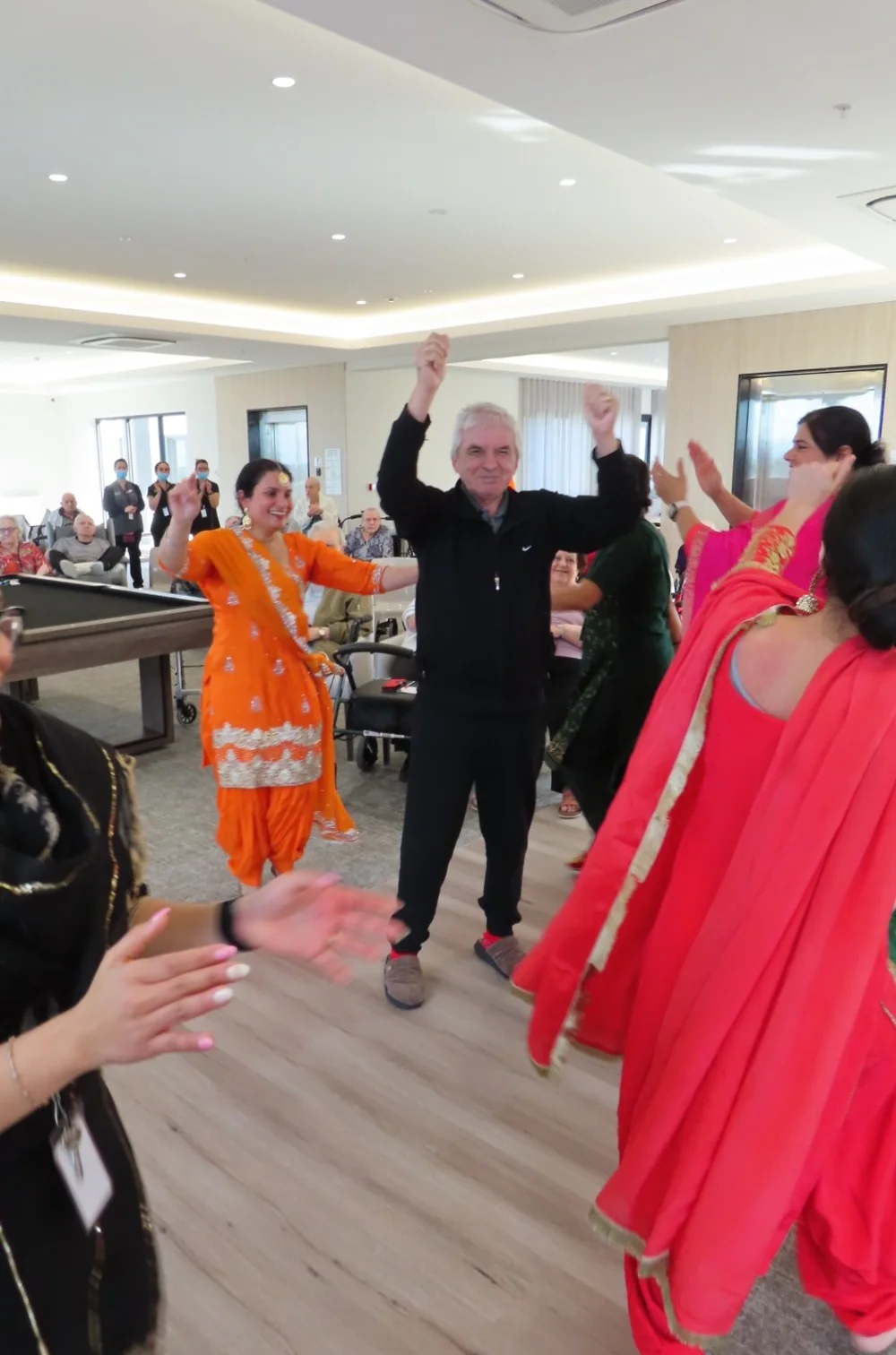 Elderly man joyfully dancing with staff and performers in colourful traditional Indian attire during a lively cultural celebration at an aged care facility.