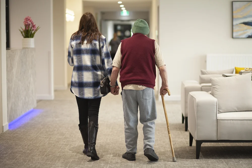 Smiling elderly Italian woman in a pink floral dress receiving support from a caregiver.