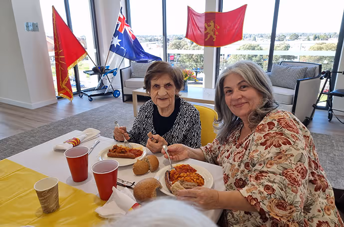 Two women enjoying a cultural meal together at an aged care facility, with flags and decorations in the background.