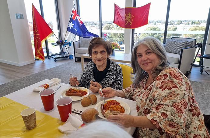 Two women enjoying a cultural meal together at an aged care facility, with flags and decorations in the background.