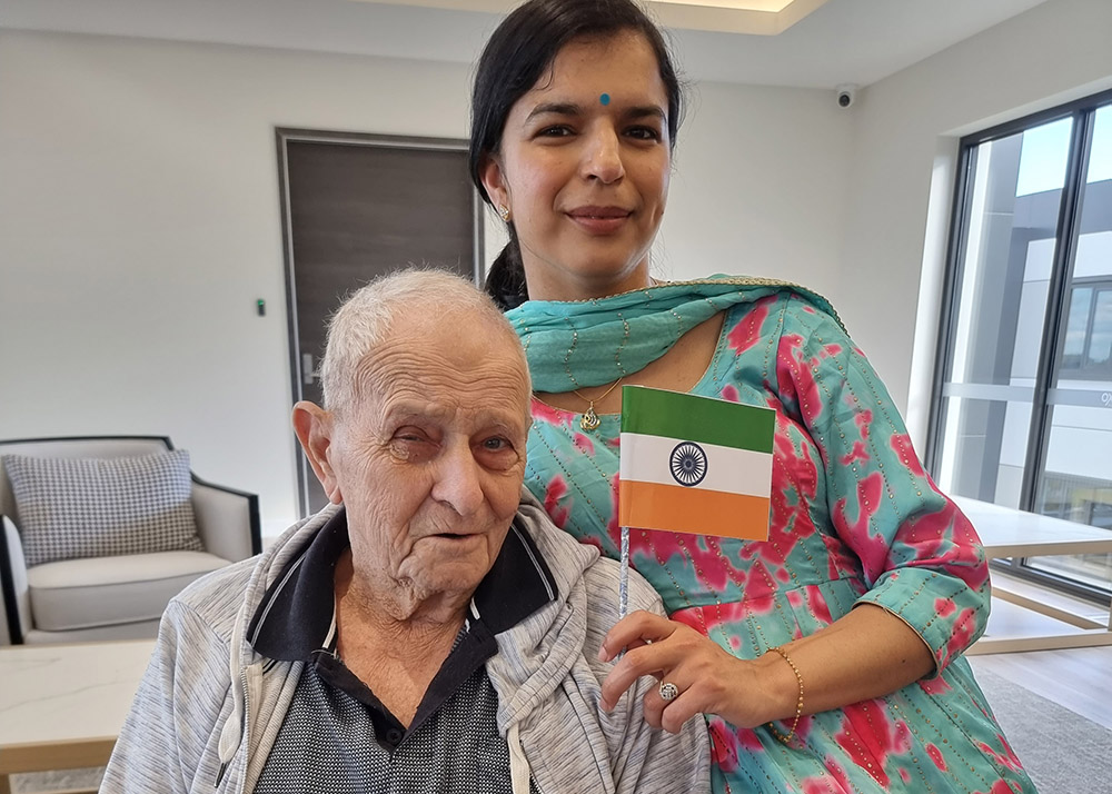 A woman in traditional dress holding an Indian flag stands beside an elderly man seated at an aged care facility.