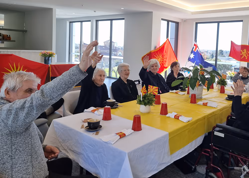 A group of elderly residents sitting around a decorated table at an aged care facility, raising their hands during a cultural celebration with flags displayed in the background.