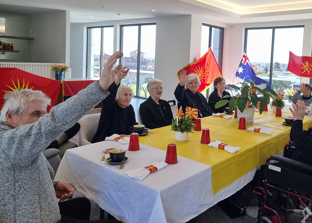 A group of elderly residents sitting around a decorated table at an aged care facility, raising their hands during a cultural celebration with flags displayed in the background.