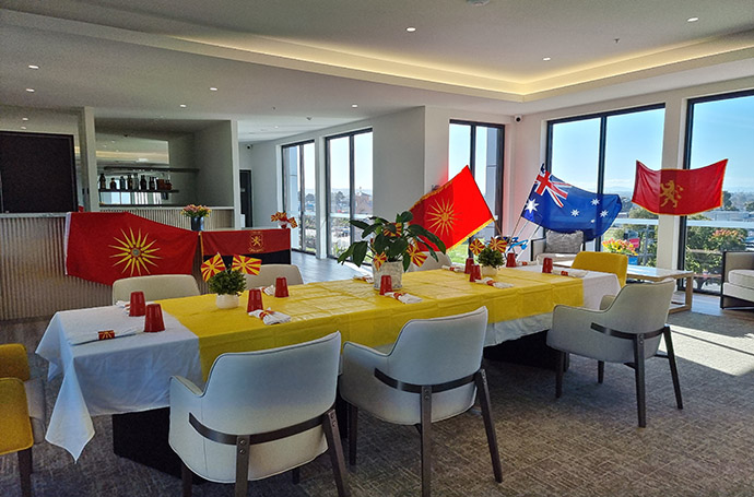 Dining area decorated with Macedonian and Australian flags, yellow tablecloths, and flowers prepared for a cultural celebration in an aged care facility.