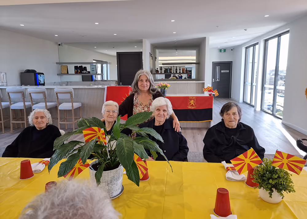 Elderly residents and a carer at an aged care facility celebrating with Macedonian flags and decorations on a yellow-themed table.]