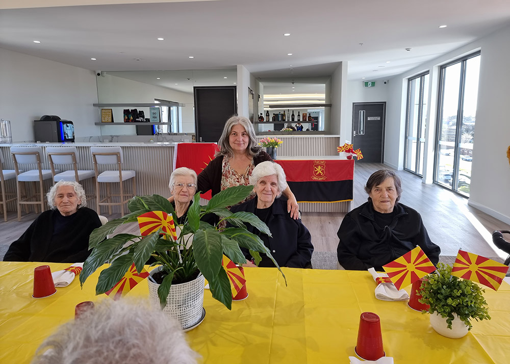 Elderly residents and a carer at an aged care facility celebrating with Macedonian flags and decorations on a yellow-themed table.]