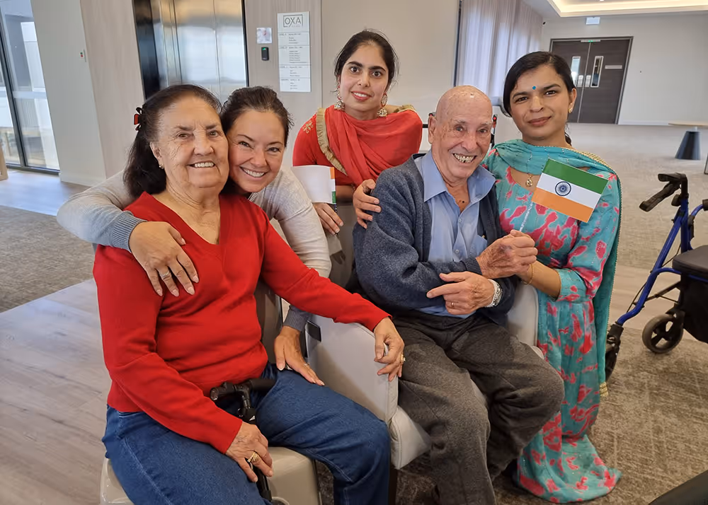 Residents and carers at an aged care facility smiling together, with one person holding the Indian flag to celebrate cultural diversity.