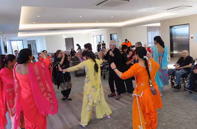 Residents and visitors in colourful traditional attire dancing together during a cultural celebration at an aged care facility.