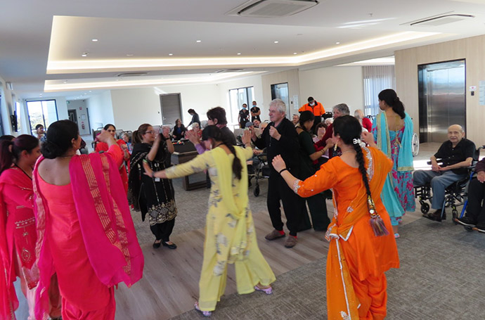 Residents and visitors in colourful traditional attire dancing together during a cultural celebration at an aged care facility.