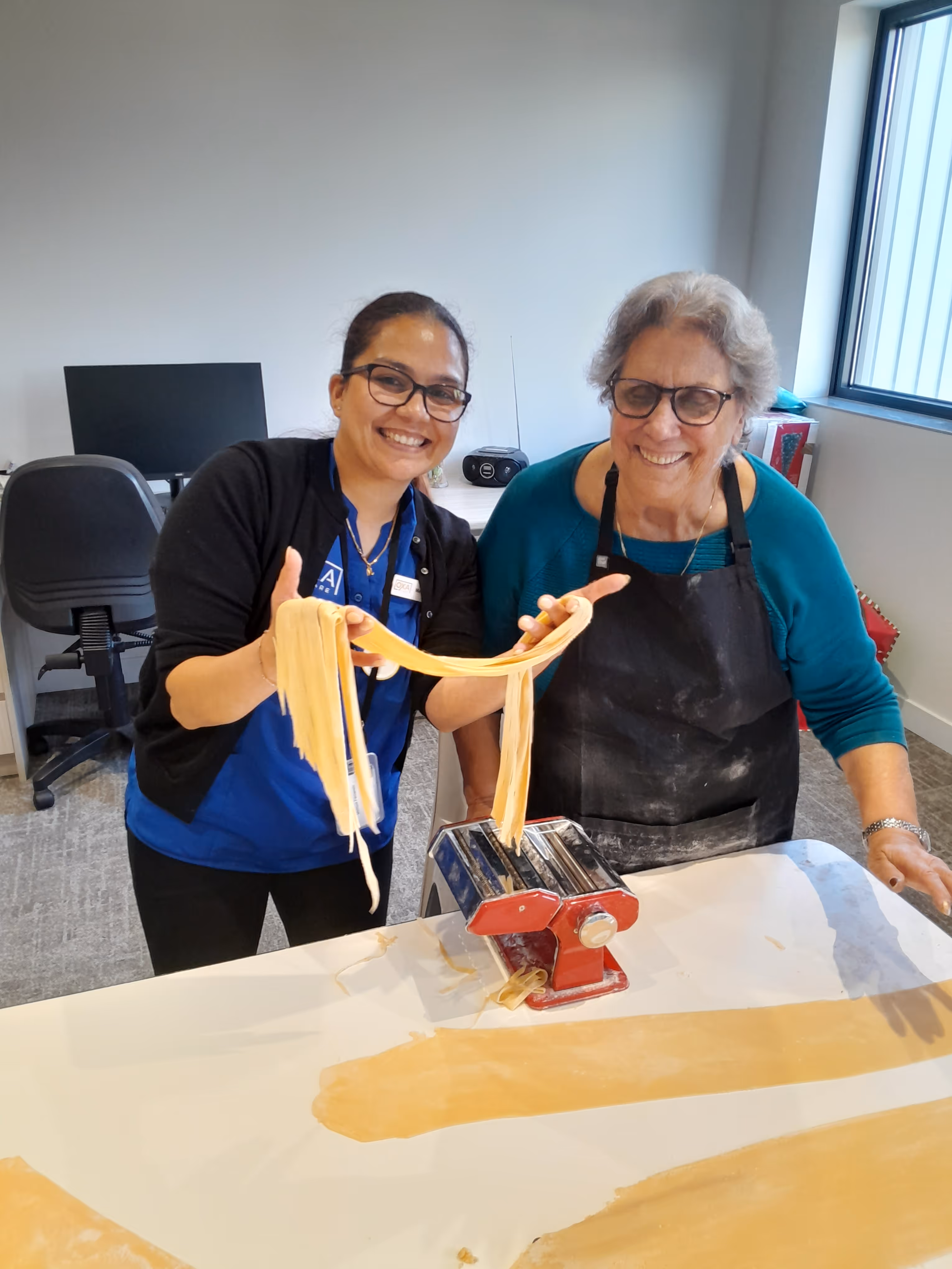 A smiling aged care resident and a staff member making fresh pasta together with a pasta machine.