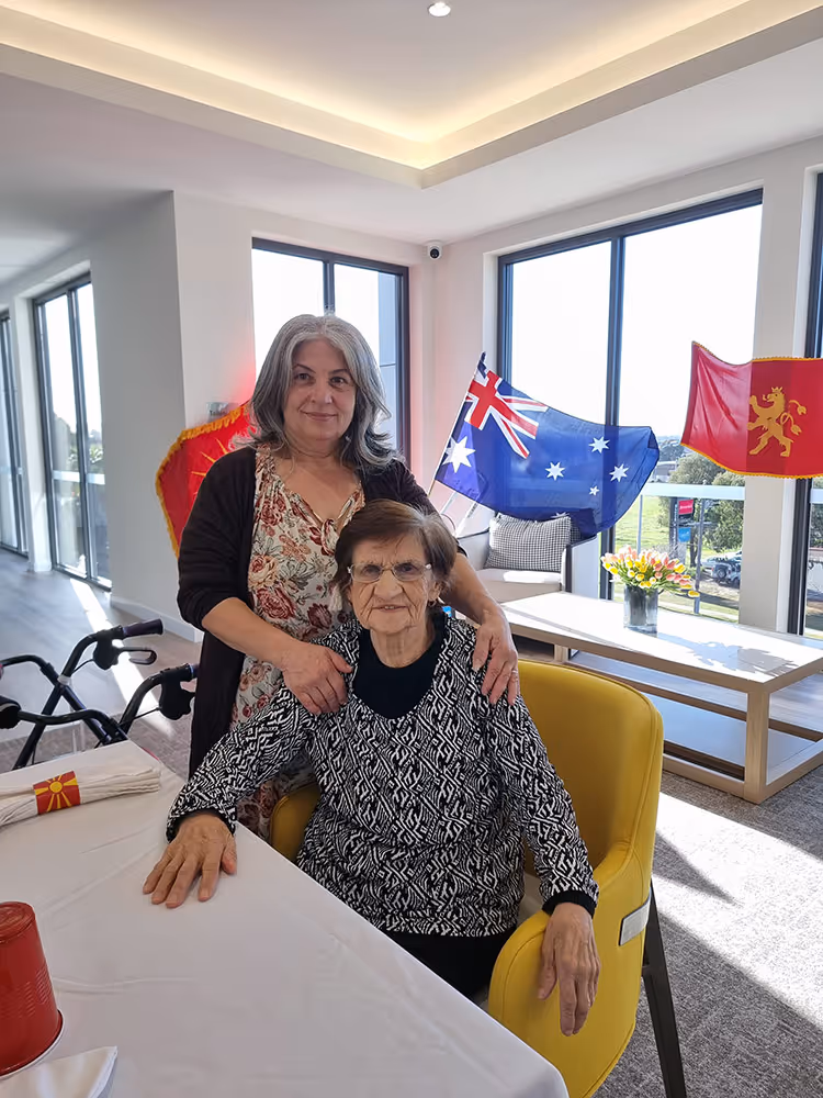An aged care resident seated at a table with a family member standing beside her, with Australian and cultural flags displayed in the background.