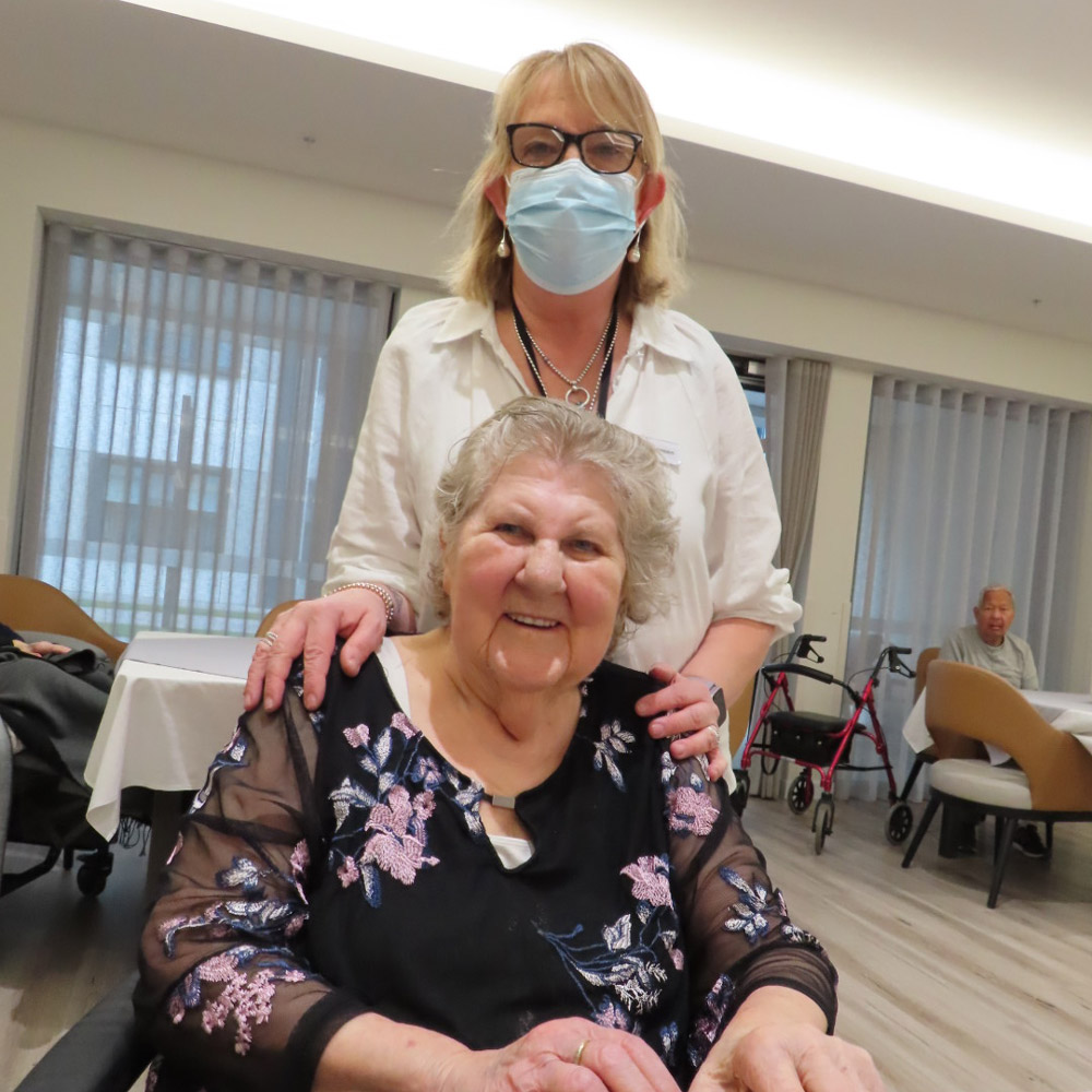 Resident seated at a table with a staff member standing beside her in a modern residential aged care home dining area.