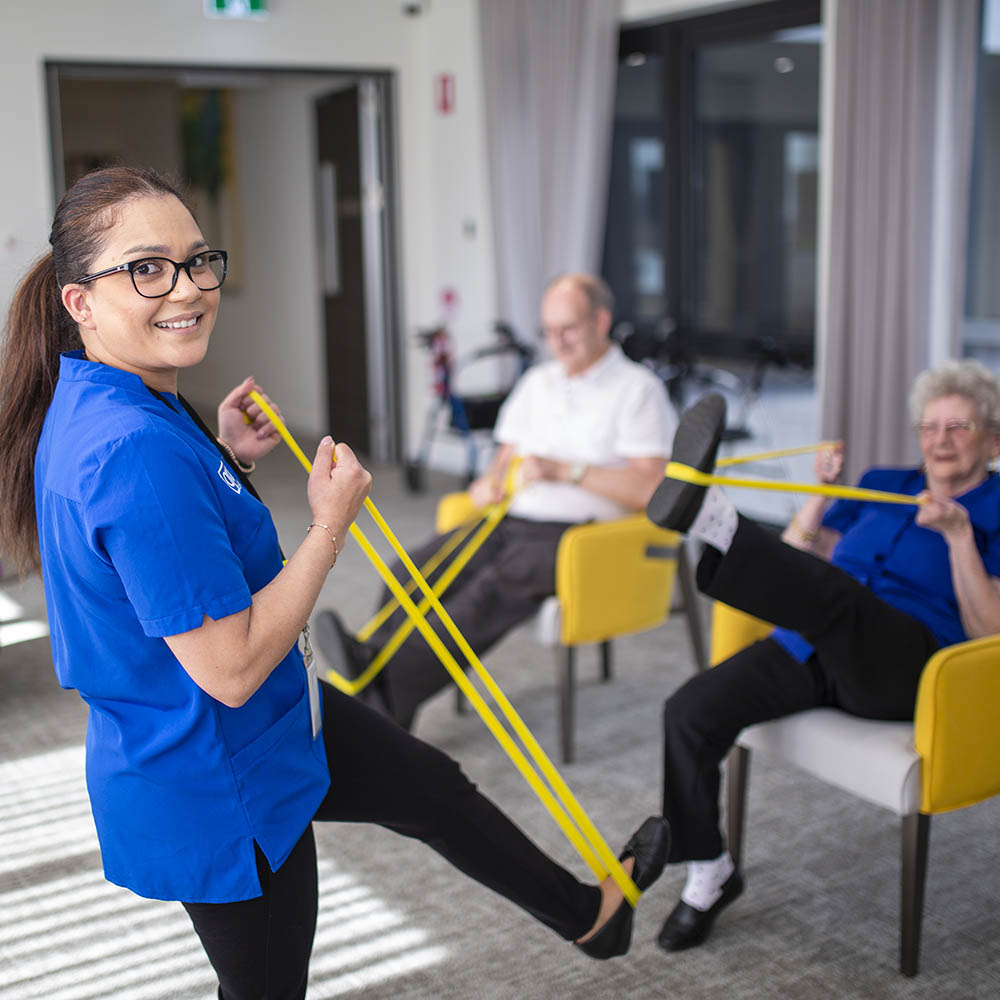Care staff leading gentle resistance band exercises with residents in a bright communal lounge area.