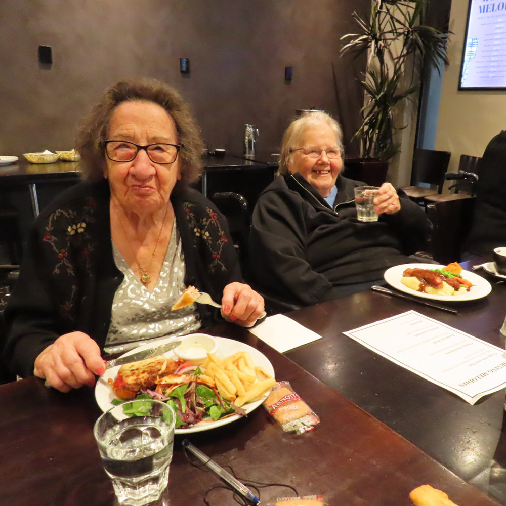 Two elderly women enjoying a meal together in a warm, social dining setting.