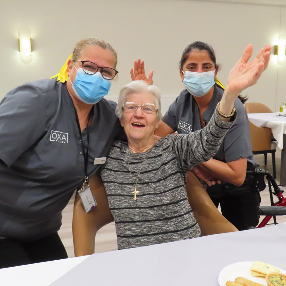 Resident celebrating with two Oxa Care team members in a bright dining area.