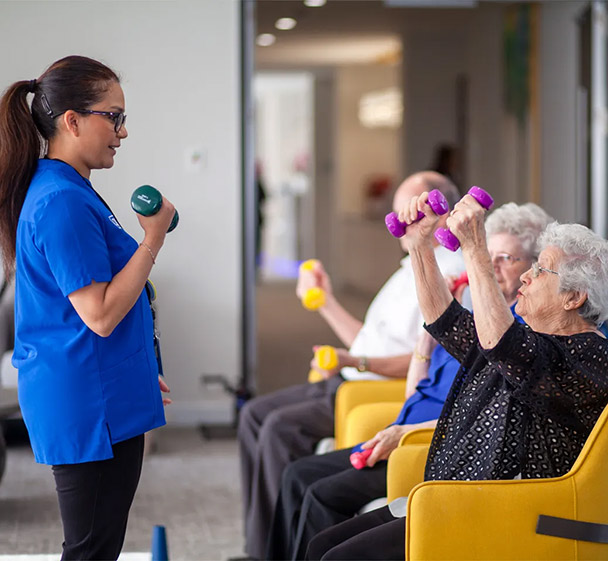 A care worker leading a gentle exercise session with elderly residents using light weights.