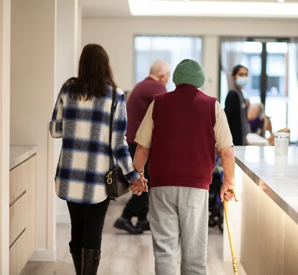 A family member walking with an elderly resident using a walking stick inside an aged care facility corridor.