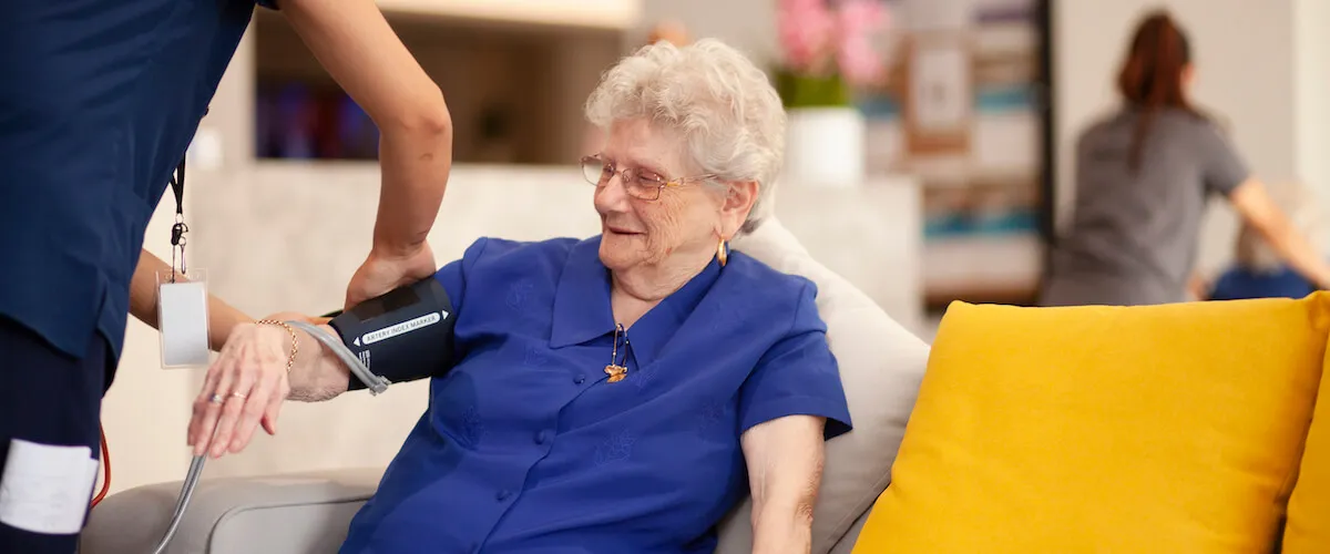 Staff member checking a resident’s blood pressure while they relax in a shared living space.