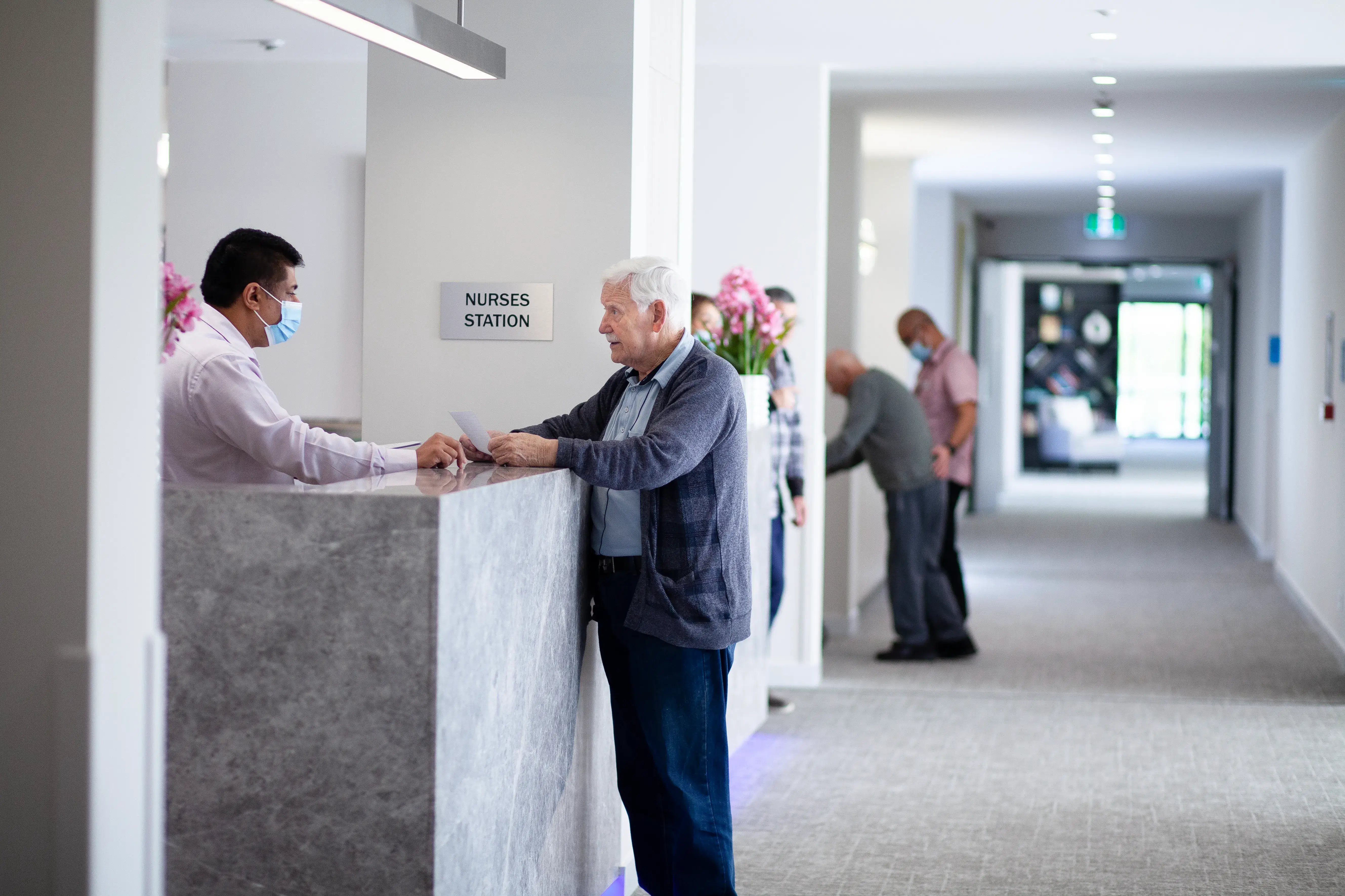 An older man speaking with a staff member at a reception counter inside a care facility.