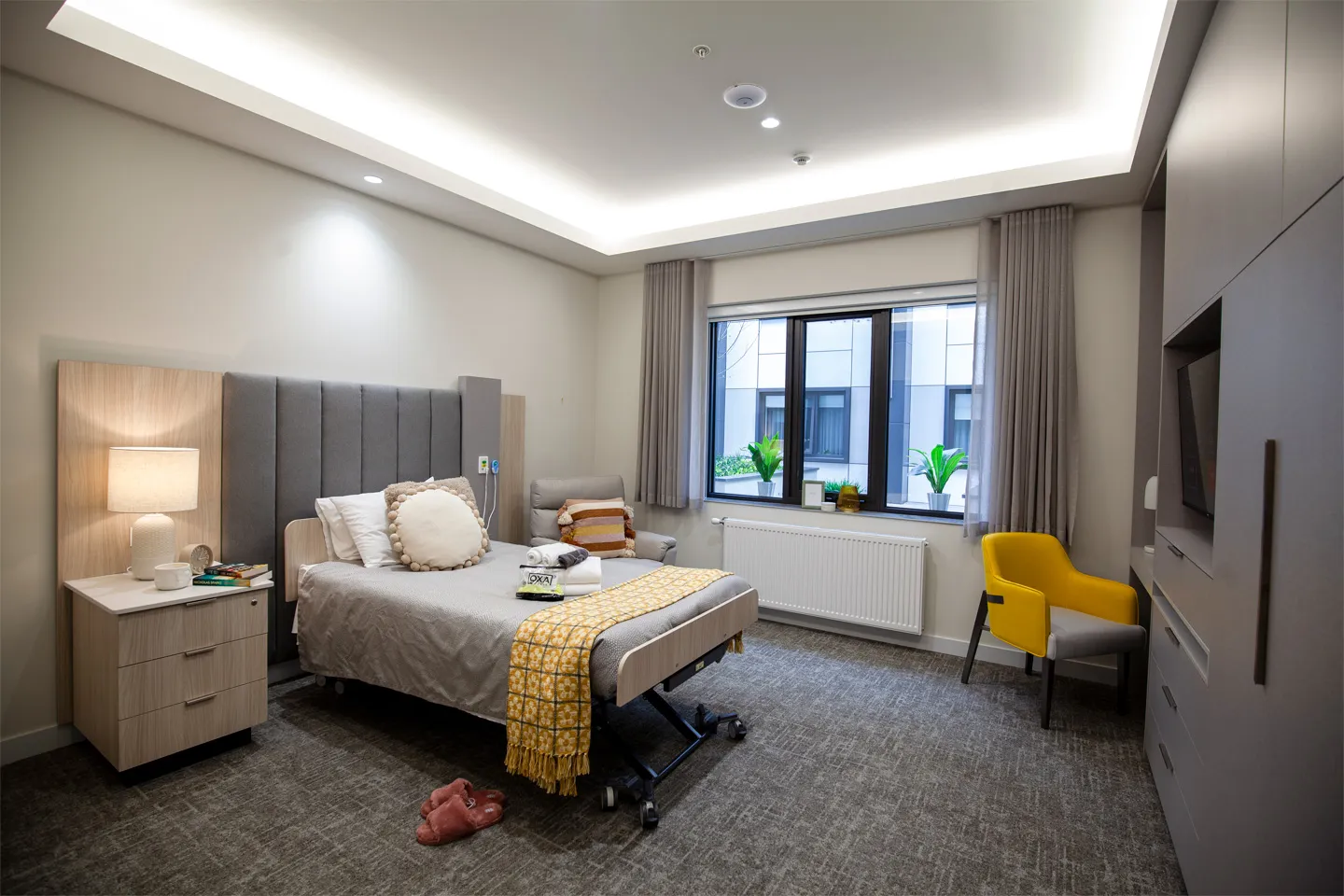 A private bedroom featuring a single bed, armchair, wardrobe, and natural light through large windows.