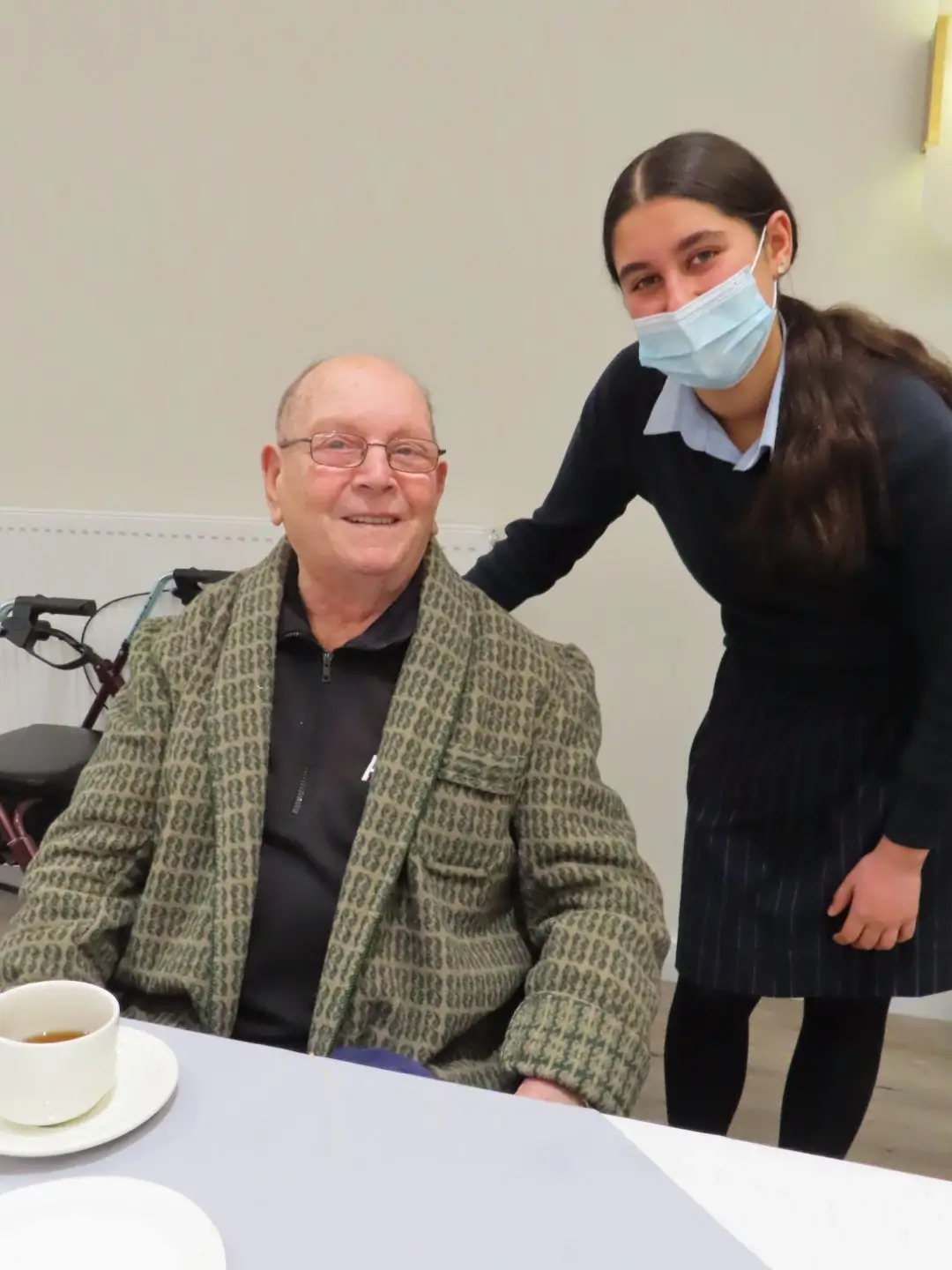  A younger support worker standing beside an older man seated at a table, both smiling during a casual visit.