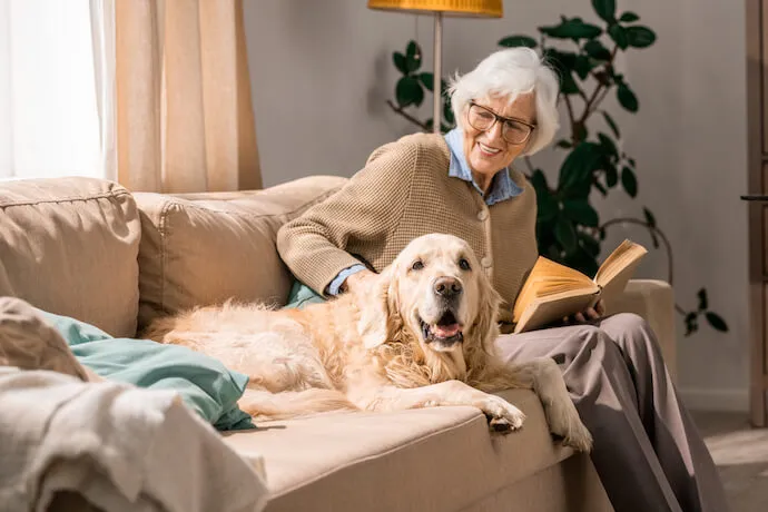 A smiling elderly resident relaxes on a sofa while reading a book and petting a golden retriever.