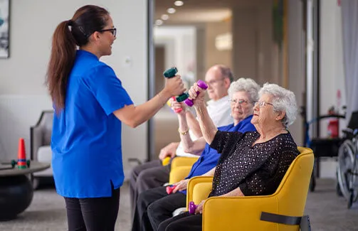 Residents perform seated dumbbell curls during a guided indoor exercise session with a staff member.