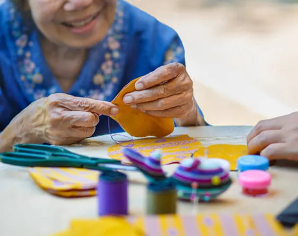 An elderly woman smiles while focusing on a colourful sewing and craft project at an Oxa Care activity session.
