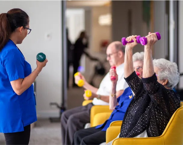 A staff member leads residents through a seated dumbbell exercise class in a bright Oxa Care wellness room.