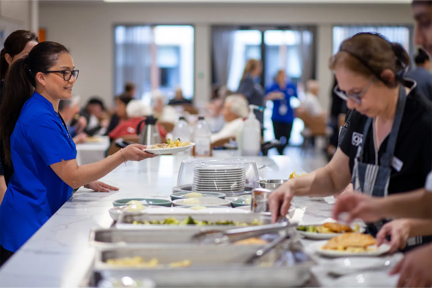 A staff member in blue scrubs serves fresh, healthy meals to residents from a modern Oxa Care serving station.