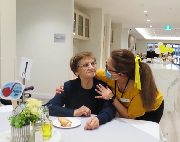 A masked staff member in yellow hugs an elderly resident during a Father's Day celebration at an Oxa Care facility.