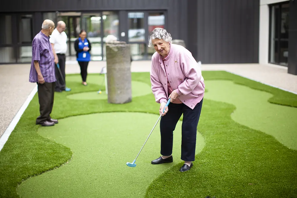 An elderly woman in a pink cardigan enjoys a game of mini-golf with friends at an outdoor Oxa Care lifestyle facility.