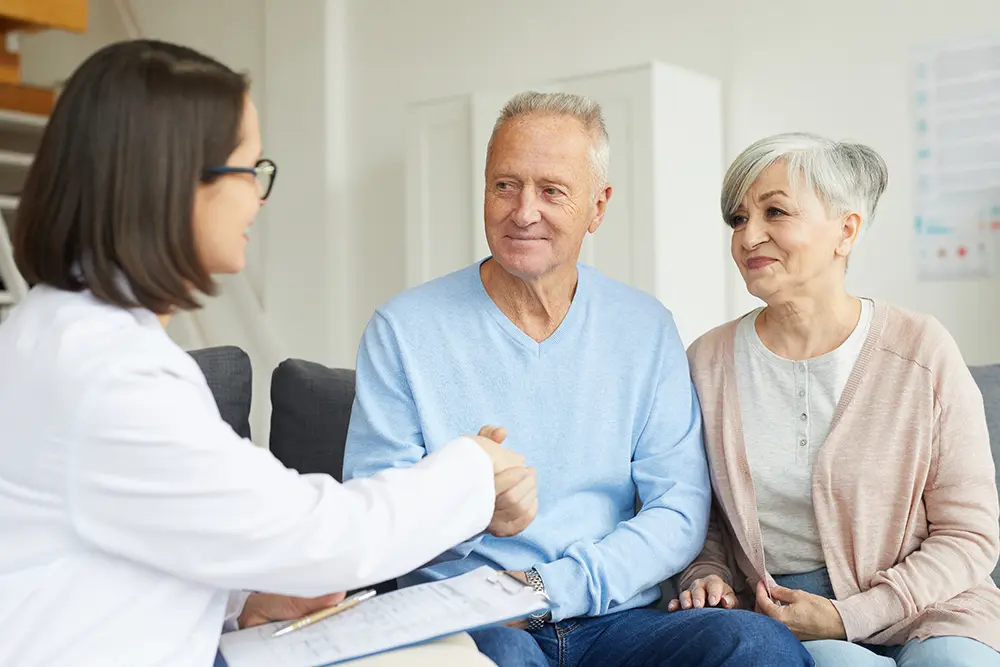 A doctor in a white coat shakes hands with an elderly couple during a supportive care consultation.