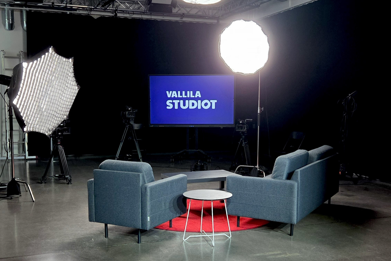 Vallila Studioiden lähetyslavastus: kaksi sohvaa, pöytä ja näyttö, kaksi kameraa jalustoilla ja studiovalot.