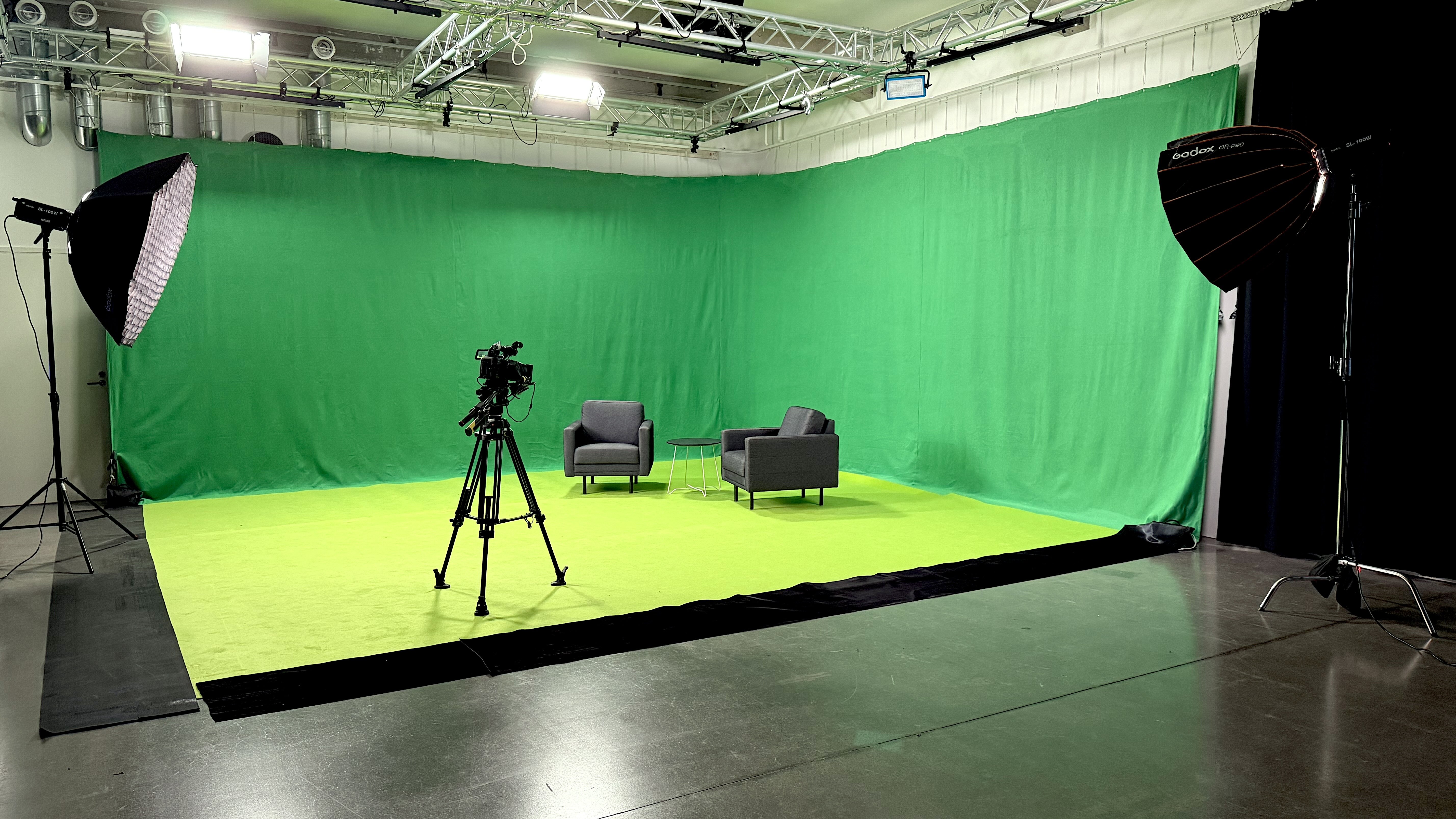 Virtuaalistudion green screen Vallila Studioiden isossa studiossa: vihreä tausta ja lattia, kaksi nojatuolia, kamera jalustalla ja studiovalot.