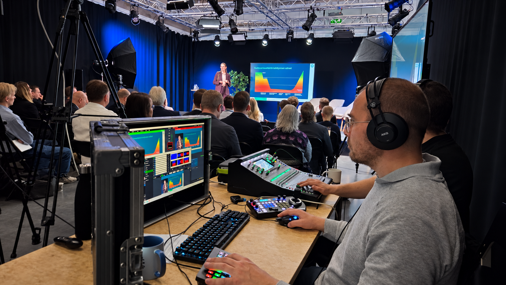 Seminaari Vallila Studioiden isossa studiossa: puhuja lavalla, yleisö tuoleilla ja etualalla tuotantopöytä, jossa teknikko ohjaa lähetystä.