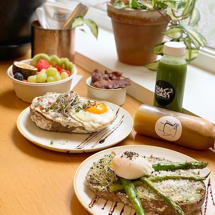 Brunch spread with poached eggs on toast, fried egg with sprouts, fresh fruit, bacon, and green juice on a wooden table by a window.