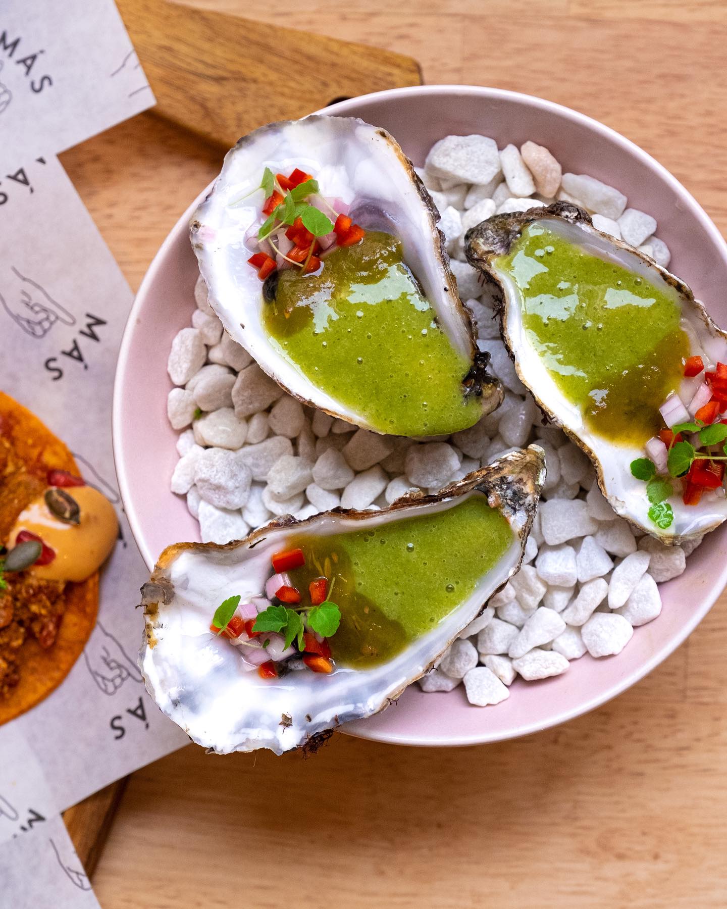 Three fresh oysters topped with green sauce and diced red peppers, served on white stones in a pink bowl.