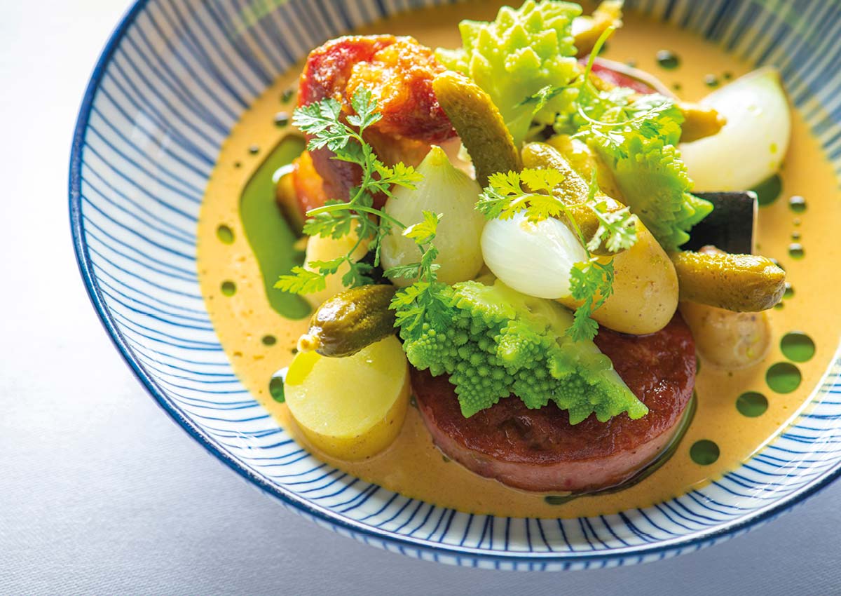 Gourmet dish in a striped bowl, featuring Romanesco broccoli, pearl onions, gherkins, and sauce over a round meat base.